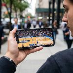 A realistic close-up of someone quickly streaming a college basketball game on their smartphone, holding the phone in one hand while on the go. The basketball game is already playing on screen, with players running on the court and a lively arena in the background of the stream.
